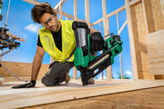 A man wearing safety glasses and a high-visibility vest kneels on a wooden deck while using a green and black Hitachi cordless nail gun.