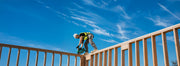 A construction worker in a high-visibility yellow vest and white hard hat is installing or adjusting a green power tool on a wooden railing outdoors under a bright blue sky with wispy clouds.